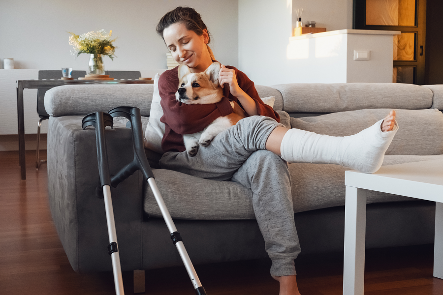 A woman sits on the sofa with her crutches beside her, wearing elasticated waistband trousers